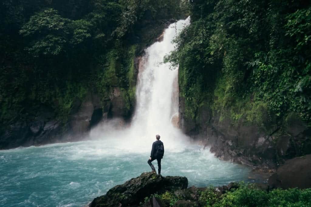 A man stands on a rocky ledge looking out at a waterfall.
