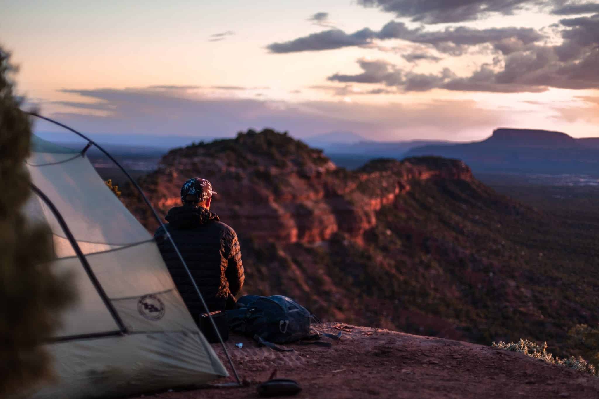 A man sitting in front of a tent looking at canyons in the distance.