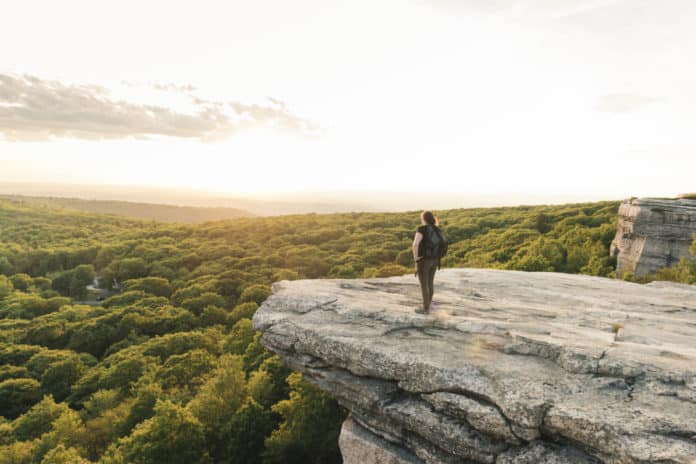 This is a horizontal, color photograph of woman hiking stopping to enjoy the view from Sam's Point Preserve in the Shawangunk Mountains. These mountains are part of the Appalachian Mountains. Green trees fill the valley below the cliffs of the mountain ridge in upstate New York, Ulster County.