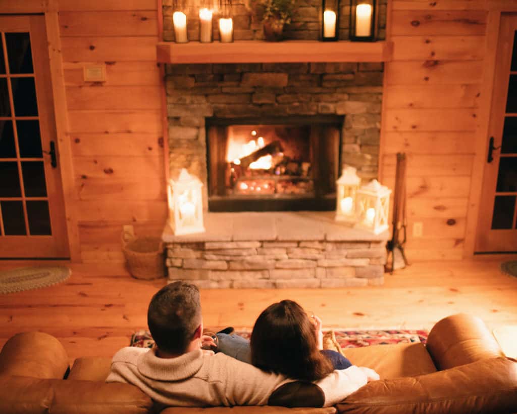 Couple sitting close together on a couch, overlooking a roaring fireplace, in a romantic warm-wood log cabin in Boone, North Carolina 