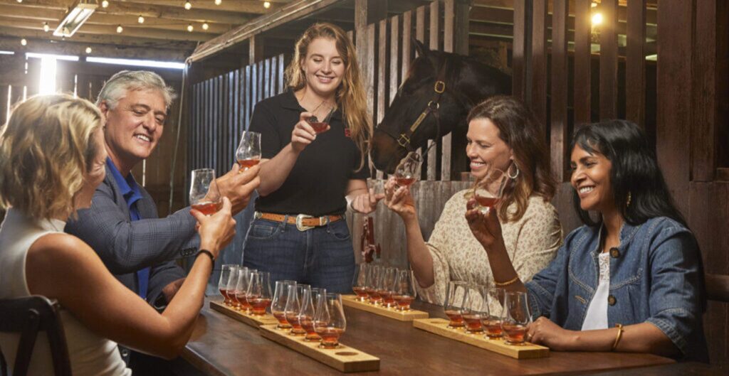 Five people sit around a wooden table in a barn, clinking glasses of whiskey or bourbon, with tasting flights in front of them. A horse stands behind them, and string lights hang from the ceiling.