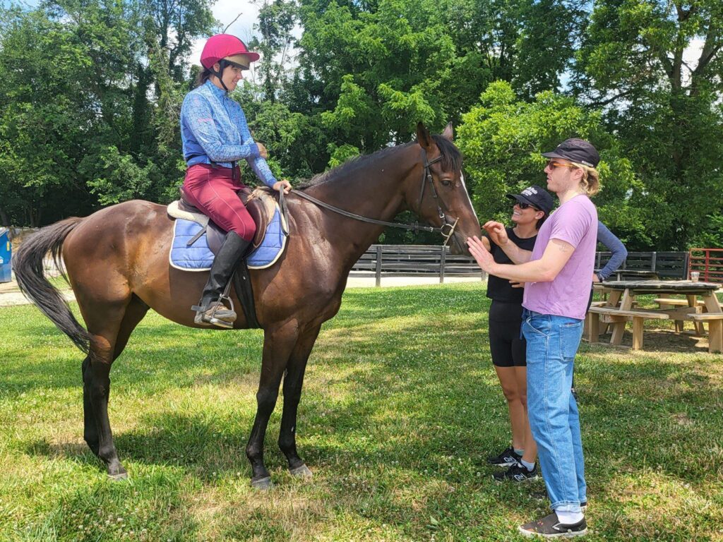 A person wearing a helmet and riding gear sits on a brown horse while two people standing beside the horse talk and gesture, with trees and a picnic table in the background on a sunny day.