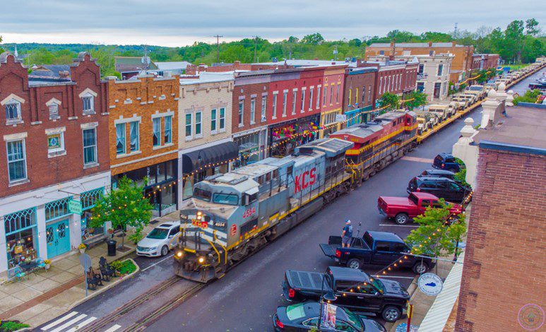 A freight train travels down tracks running through the middle of a small town’s Main Street lined with colorful historic buildings, shops, parked cars, and people on a cloudy day.
