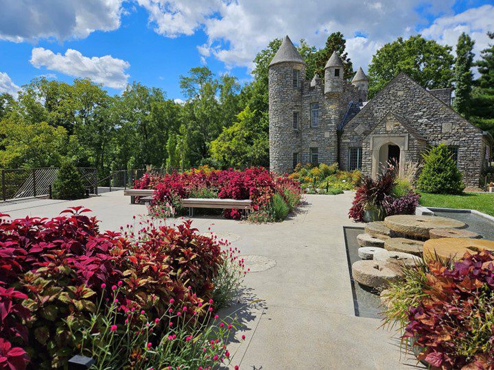 A stone castle-like building with round towers sits behind vibrant red and green gardens and a paved courtyard under a partly cloudy blue sky. Benches and a stone water feature are visible in the foreground.