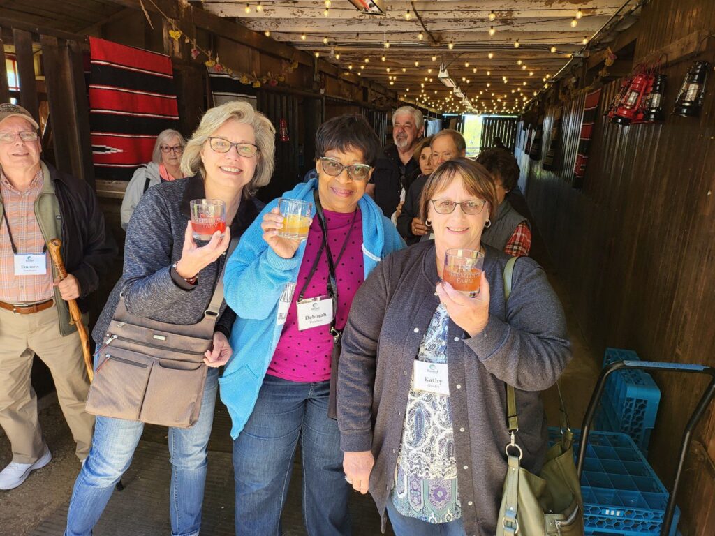 A group of smiling adults stand in a rustic, string-lit hallway, holding drinks and wearing name tags. Three women in front pose closely together, while others gather behind them, enjoying the cheerful atmosphere.