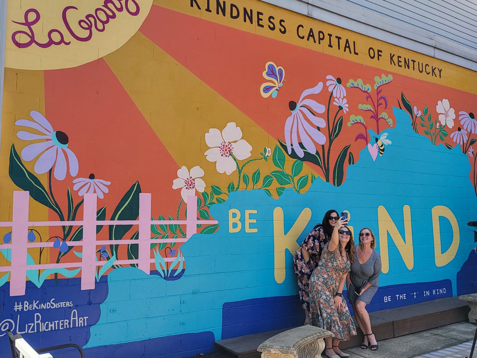 Three women smile and pose together in front of a colorful mural that says Be Kind with flowers, sun rays, and “Kindness Capital of Kentucky” written at the top. The mural promotes kindness and positivity.