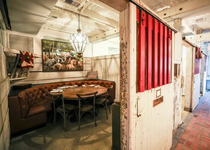 A cozy restaurant booth with brown tufted seating and a round wooden table, set inside a rustic space with white wooden walls, red accents, and a barn-like decor theme. Brick flooring and farm-themed artwork are visible.