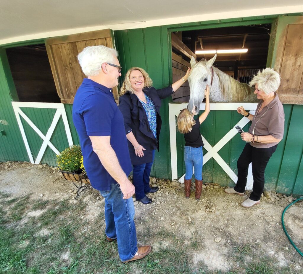 Four adults and a young girl stand by a green barn. The girl pets a white horse sticking its head out of a stall window while the adults smile and watch. There is a pot of yellow flowers and a garden hose nearby.