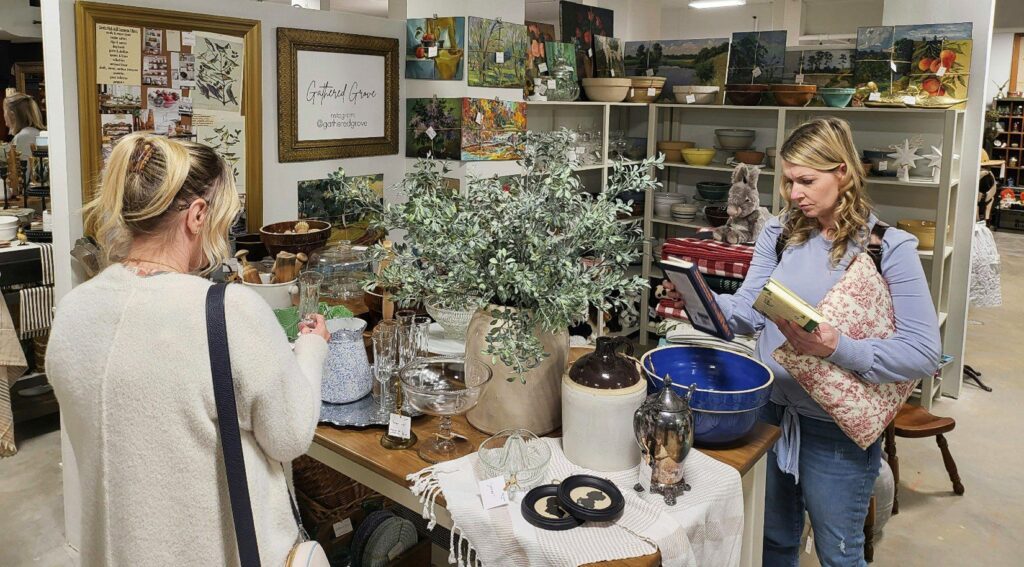 Two women browse items at an antique store, examining dishes, bowls, and decor on tables and shelves. The store is filled with various vintage items and artwork. One woman holds a book and inspects an object.