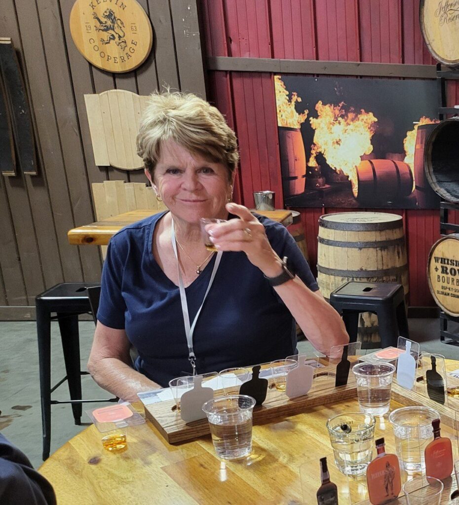 A woman with short brown hair sits at a table holding a tasting glass, smiling. She is at a distillery, with barrels, tasting boards, and a fiery display in the background.