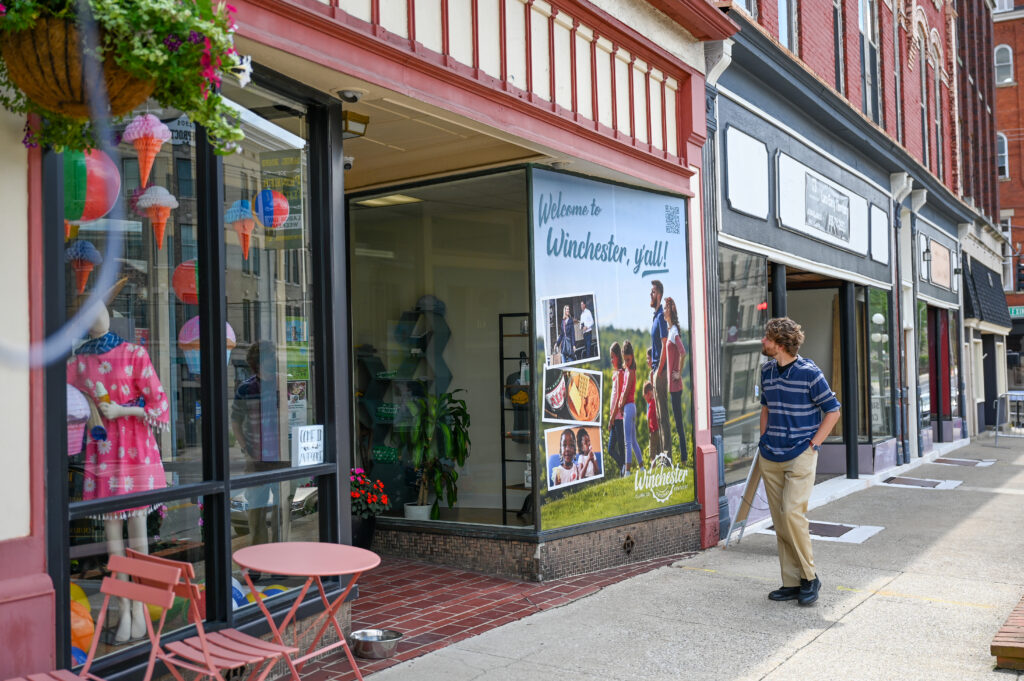 A man in a striped shirt and tan pants walks along a sidewalk past a shop window displaying a “Welcome to Winchester, y’all!” sign with photos of people and landscapes.