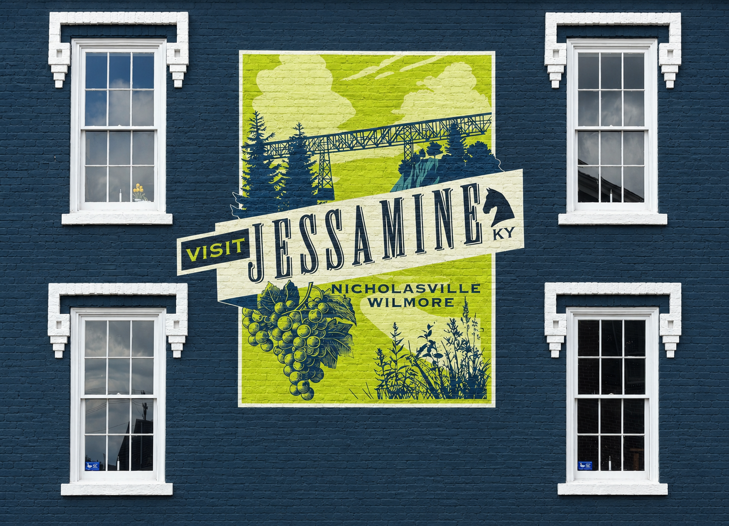 A mural on a blue brick wall reads “Visit Jessamine KY, Nicholasville Wilmore” with images of a bridge, trees, and grapevines between four white-trimmed windows.