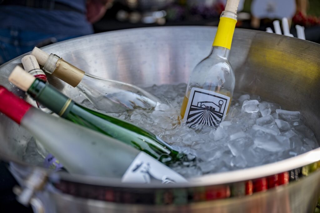 Several wine bottles chill in a large metal tub filled with ice. One bottle with a yellow neck and a train label is most prominent in the foreground.