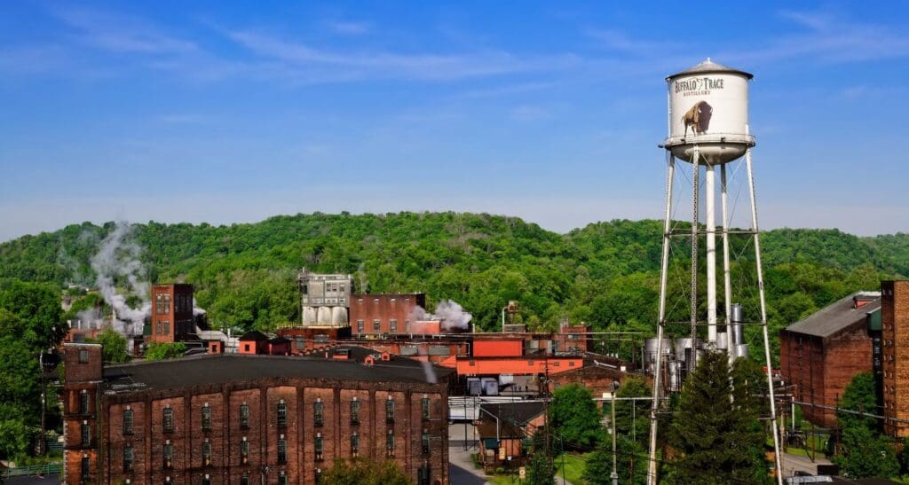 A distillery complex with red brick buildings and a tall white water tower labeled Buffalo Trace. Smoke rises from chimneys. Lush green trees and a clear blue sky form the backdrop.