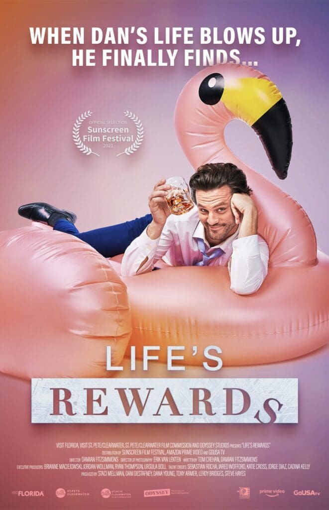 A man in a suit, holding a glass, sits inside an inflatable pink flamingo with blue water in the background. Text above reads, When Dans life blows up, he finally finds... The title Lifes Rewards is printed below. Film festival laurels are visible.