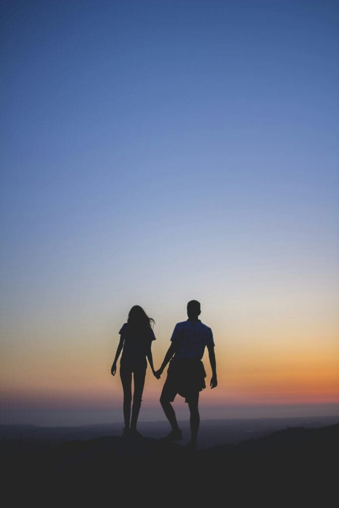 Silhouette of a couple holding hands, standing on a hill at sunset with a colorful sky gradient from deep blue to orange.