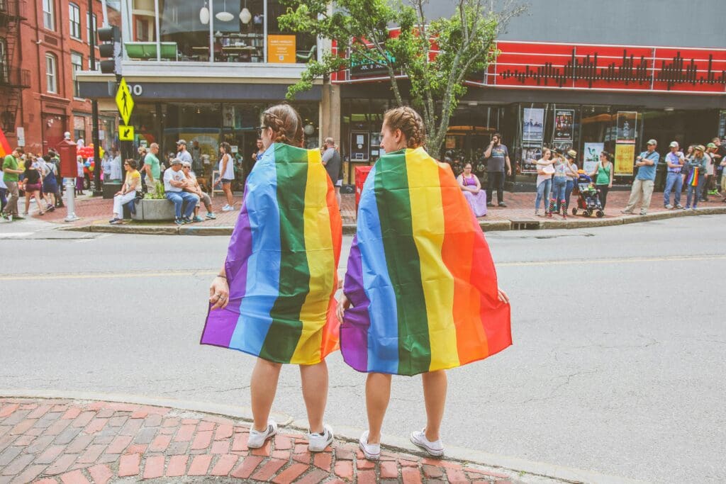 Two people stand on a sidewalk, draped in rainbow flags, symbolizing LGBTQ+ pride. They face a street, watching a crowd across the road. The setting is a bright, urban environment with spectators lining the opposite sidewalk.