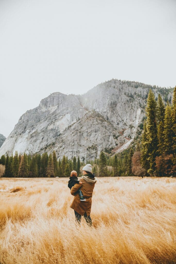 A person wearing a coat and hat holds a baby in a field of tall, golden grass. In the background, there are tall evergreen trees and a large rocky mountain under a pale sky.