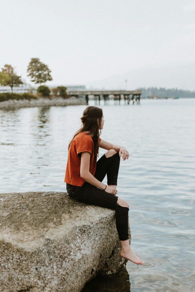 A person with long hair and a red shirt sits on a large rock by the water, looking out at the horizon. Barefoot, they rest one arm on their knee. A pier and trees are visible in the background.