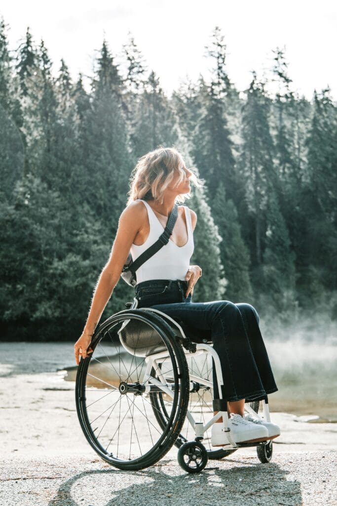 A woman with blonde hair sits in a wheelchair outdoors, wearing a white top and dark pants. She is surrounded by trees and sunlight filters through the forest. She looks to the side, with one hand resting on her wheel.