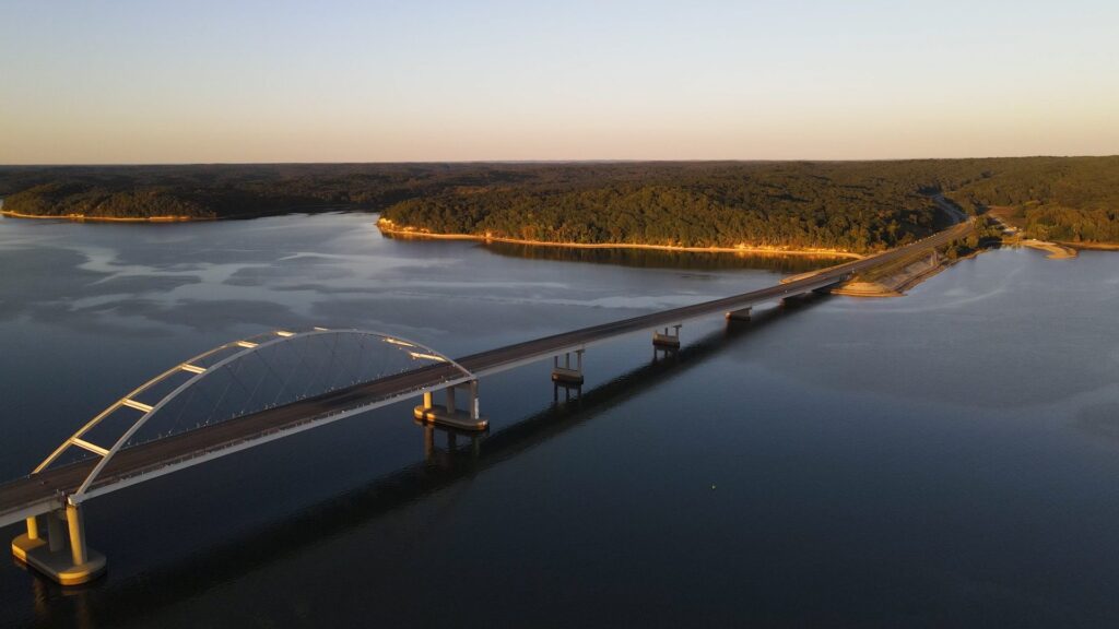 A long bridge with an arched section spans across a calm body of water at sunset, surrounded by dense green forest on the shoreline.