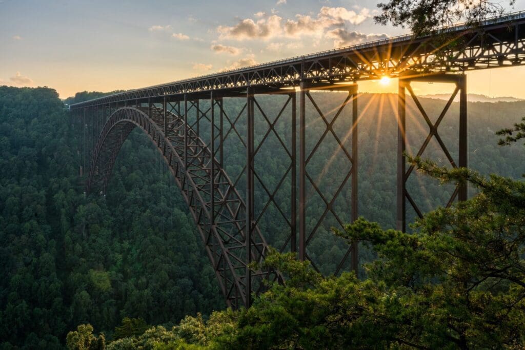 The image shows a large steel arch bridge spanning a lush, green forest. The sun is setting, casting a warm glow and sun rays through the bridges structure. Fluffy clouds accent the sky, with trees partially framing the scene.