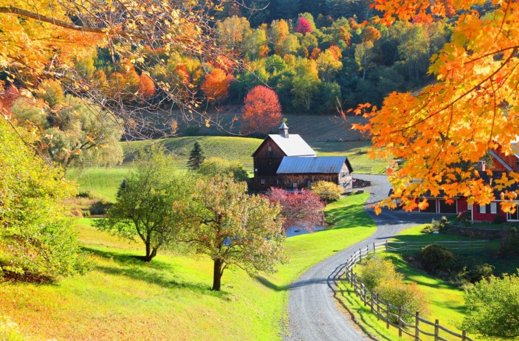 A scenic autumn landscape featuring a rustic barn surrounded by vibrant fall foliage in shades of orange, red, and yellow. A curving gravel road leads to the barn, with lush green grass and trees lining the path.