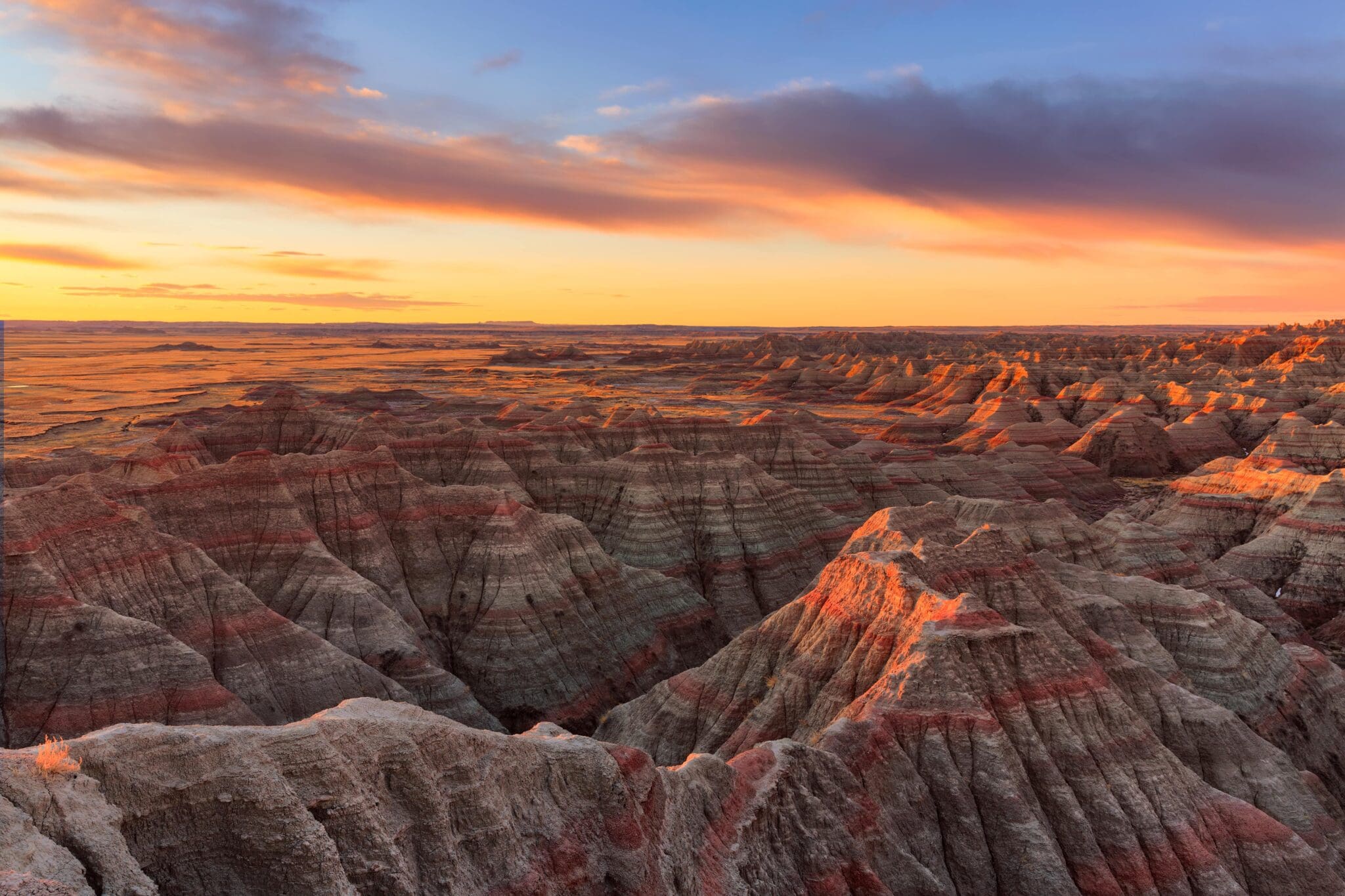 Sunrise over the Badlands National Park, featuring colorful, striped rock formations and rugged canyons. The sky is painted with soft oranges and blues as clouds stretch across the horizon.