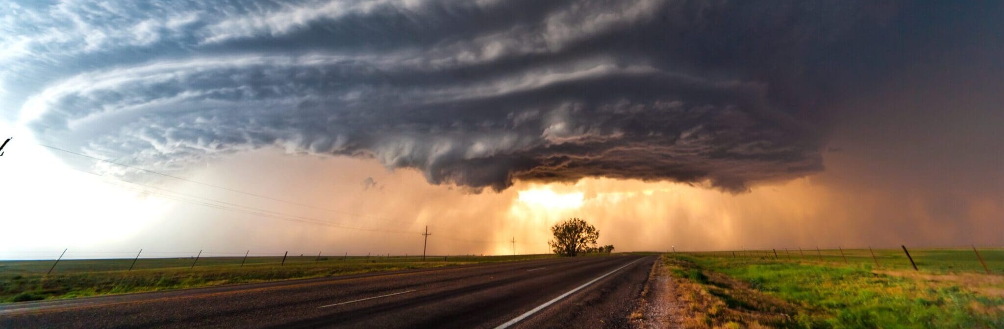 A dramatic storm cloud formation looms over an open, deserted road cutting through a grassy landscape. The swirling clouds are dark and ominous, with a hint of sunlight breaking through, casting a dramatic light on the scene.