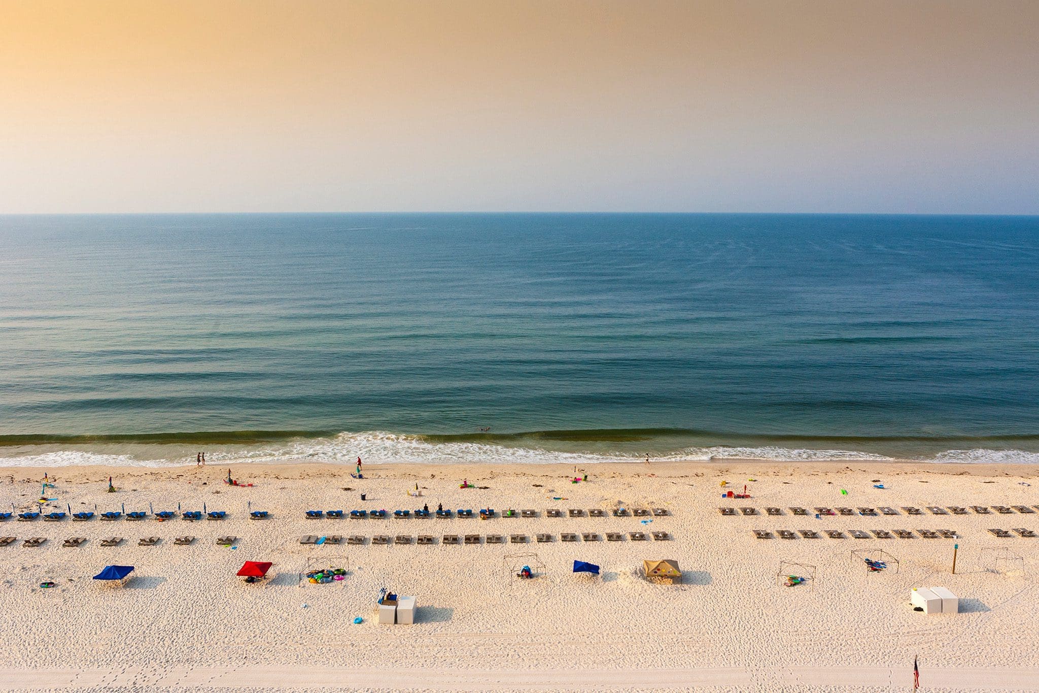 Aerial view of a sandy beach lined with empty sun loungers and umbrellas. The calm ocean stretches out to the horizon under a clear, hazy sky. A few people are scattered on the beach and in the water.