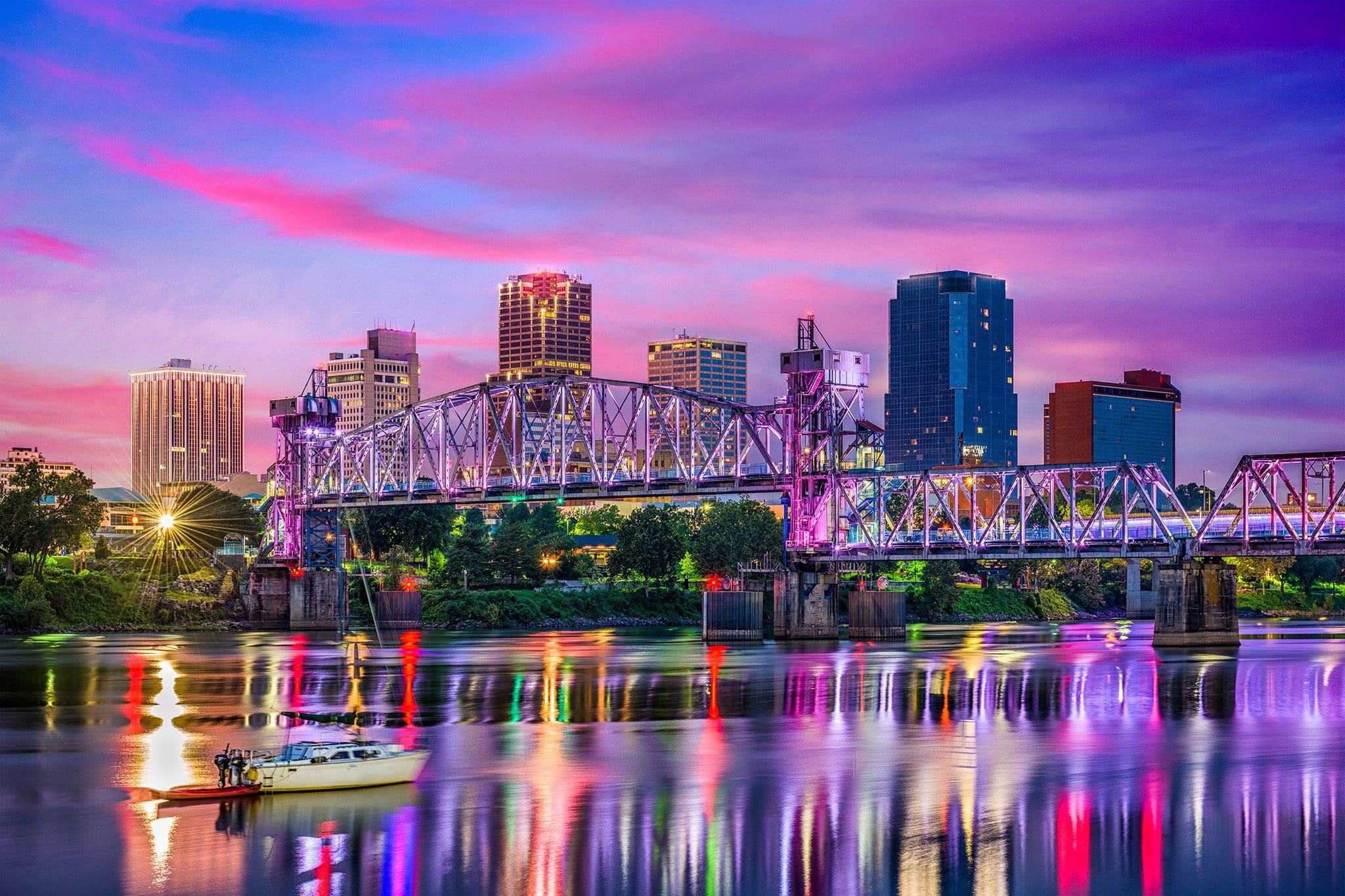 A vibrant cityscape at dusk featuring a lit-up bridge spanning across a calm river. The skyline is dotted with colorful, illuminated buildings against a pink and purple sky. A small boat floats in the water, reflecting the citys lights.