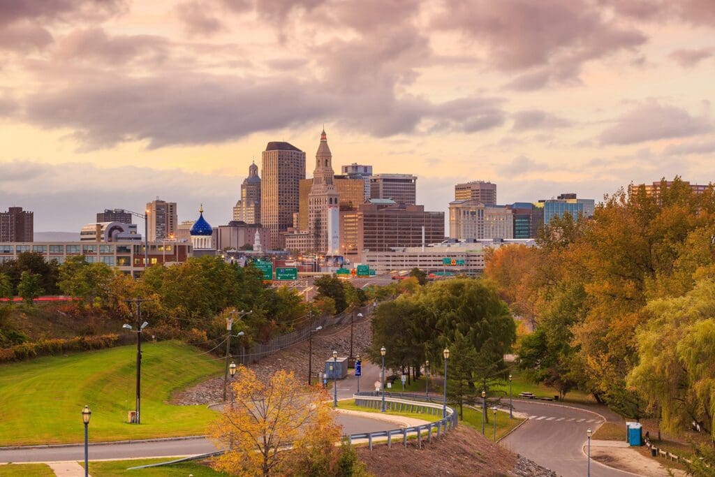 A city skyline under a cloudy sky at sunset, featuring tall buildings and a domed structure. In the foreground, theres a park with a winding path, lamp posts, and autumn trees with colorful leaves.