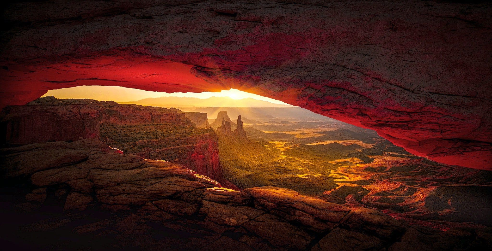 Sunrise view through a rocky arch, illuminating a canyon with warm light. Rugged terrain and rock formations are visible under the glowing sky, creating a dramatic and picturesque landscape.
