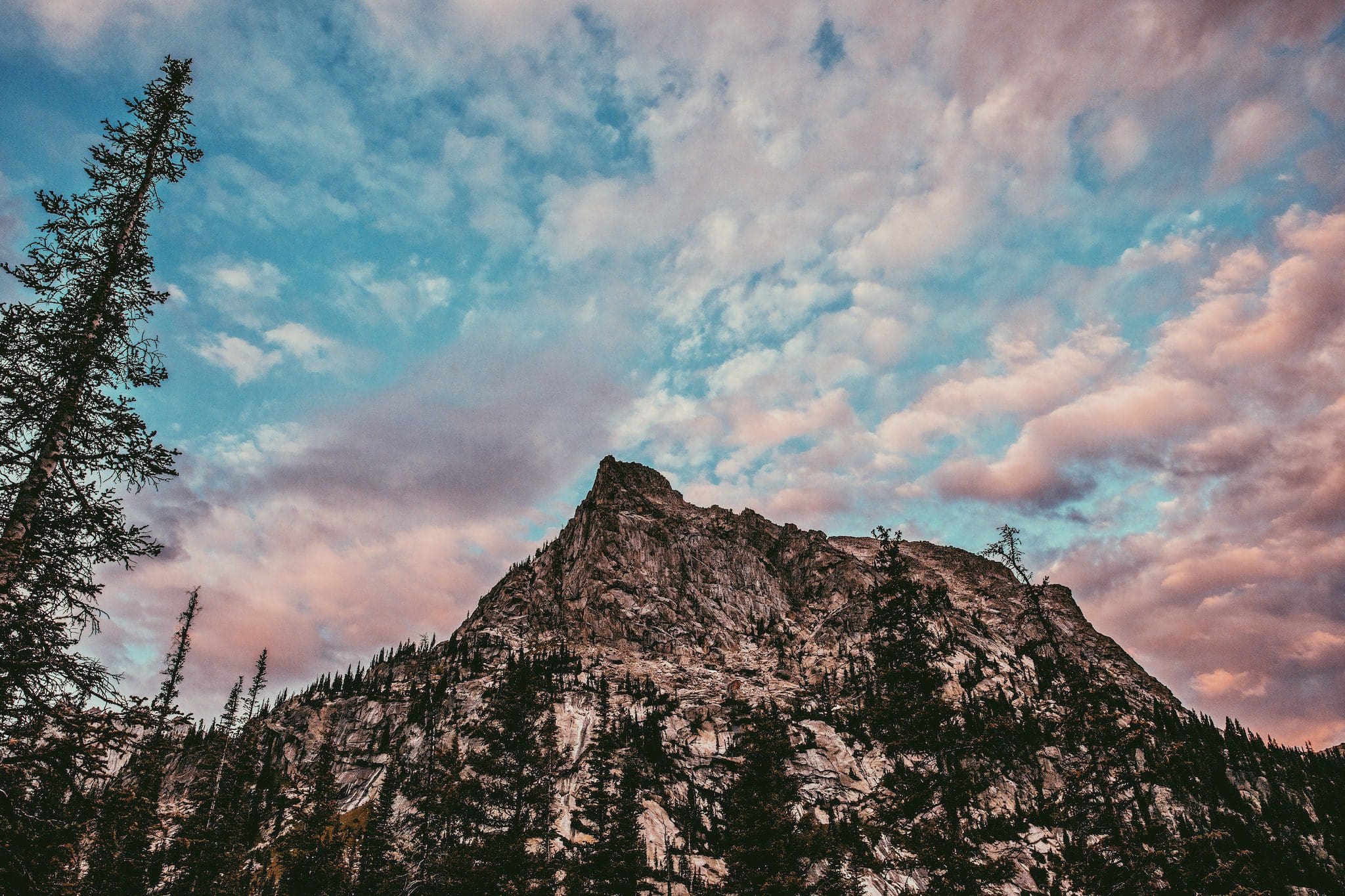 Mountain peak surrounded by pine trees under a sky filled with pink and blue clouds at sunset. The rocky terrain contrasts with the vibrant, colorful sky, creating a dramatic scene.