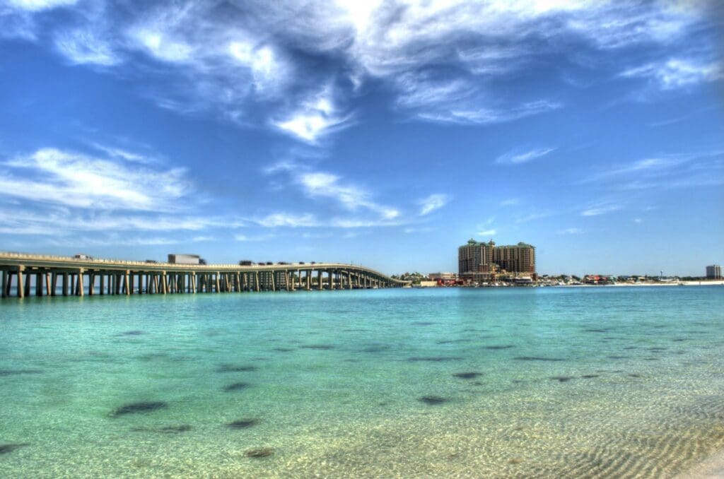 A serene ocean scene with clear turquoise water in the foreground. A long bridge stretches across the water to the left, leading to a coastal city with tall buildings under a partly cloudy blue sky.