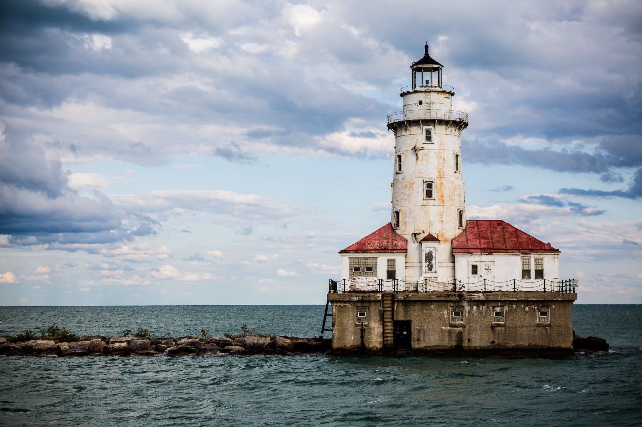 A solitary lighthouse with a red roof stands on a rocky foundation amidst a body of water. The sky above is cloudy with patches of blue. Waves gently surround the structure, conveying a sense of isolation and tranquility.
