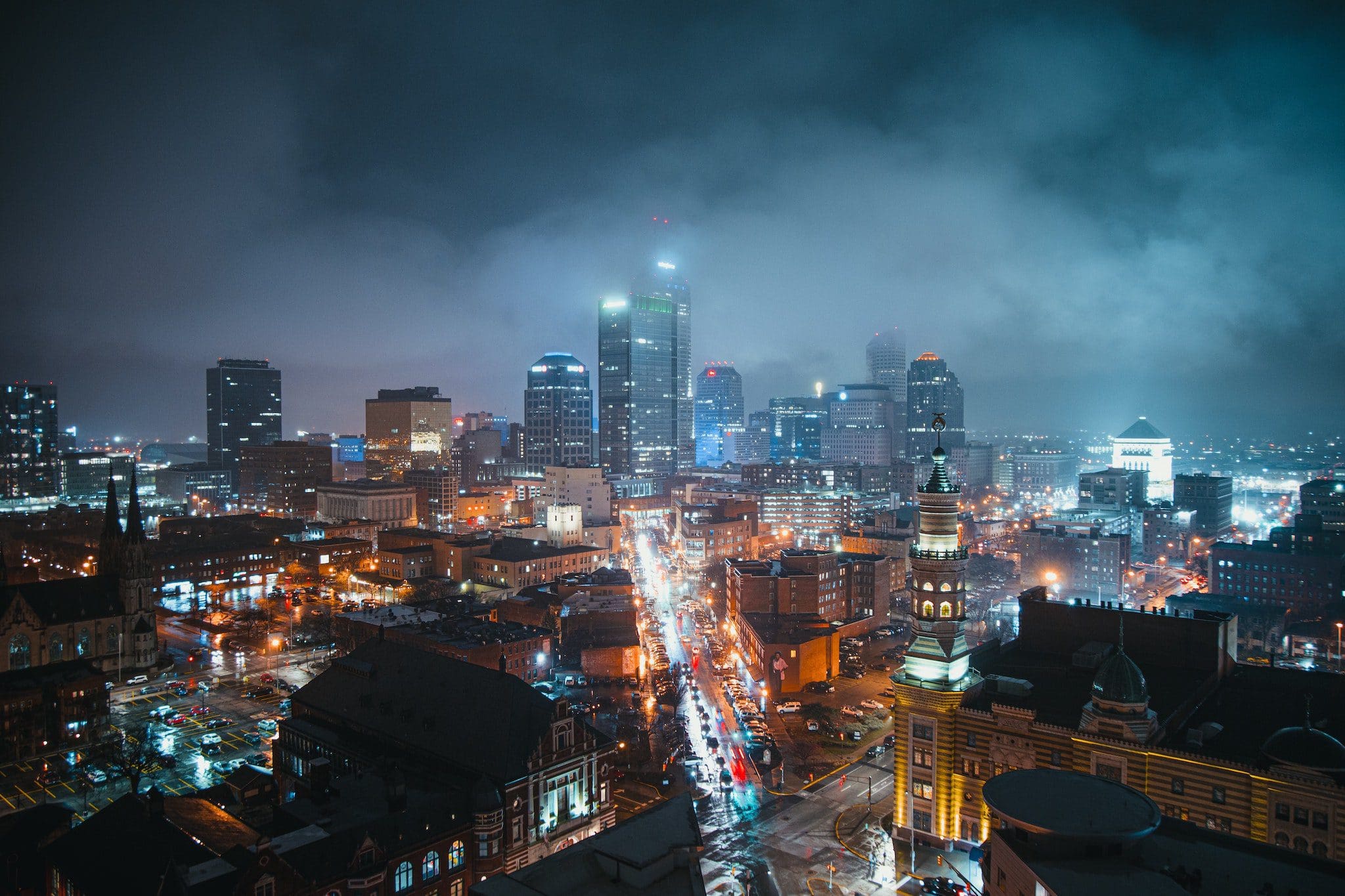 A nighttime cityscape with a bustling street lined by illuminated buildings. Skyscrapers with glowing windows are visible through a haze. The streets are busy with car lights, and a cloudy sky hovers above the city.