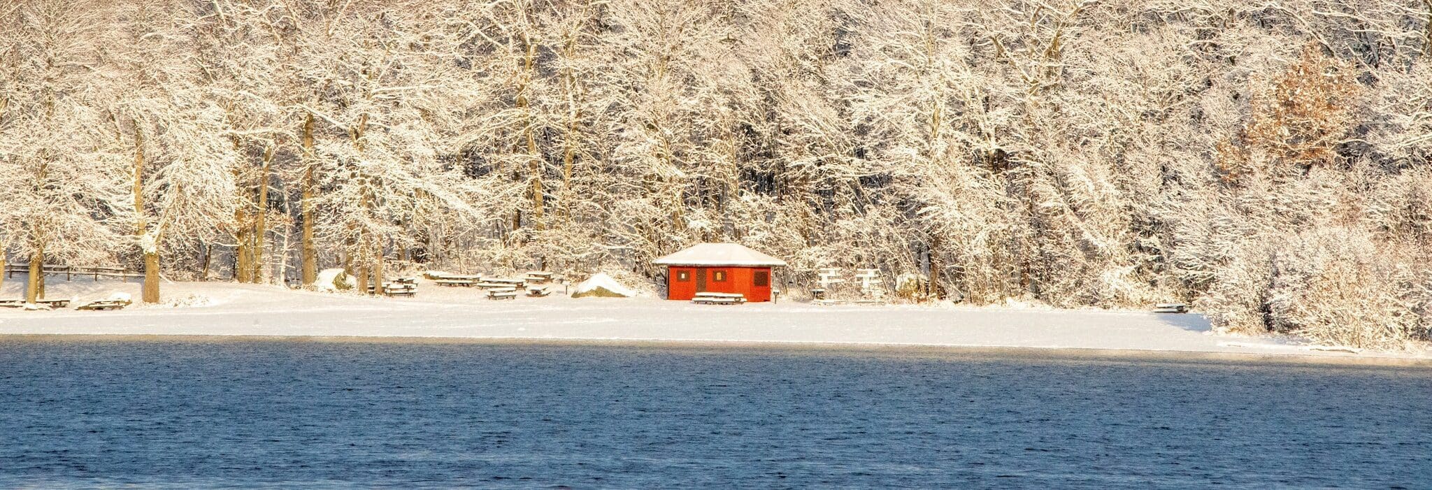 A small red cabin is nestled among snow-covered trees on the shoreline of a lake. The icy blue water contrasts with the snowy landscape, creating a peaceful winter scene.