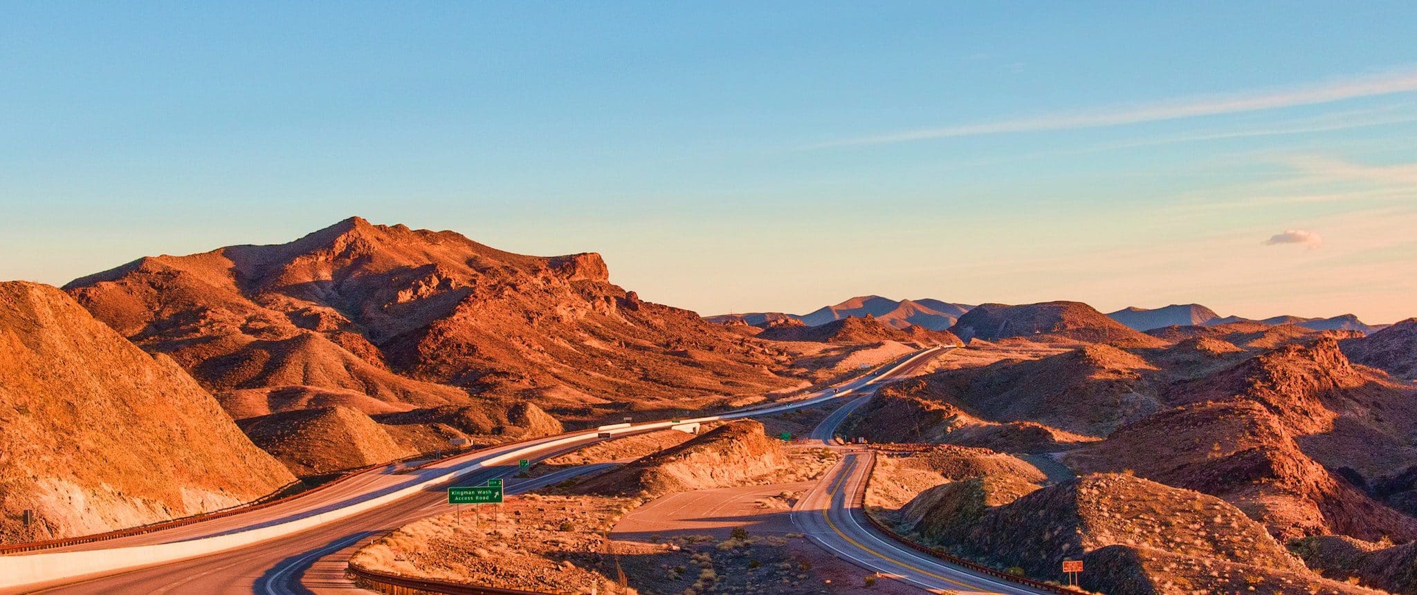 A scenic view of a highway winding through a rocky desert landscape at sunset. The sky is clear with a warm glow, highlighting the rugged terrain and distant mountain peaks. A green road sign is visible along the road.