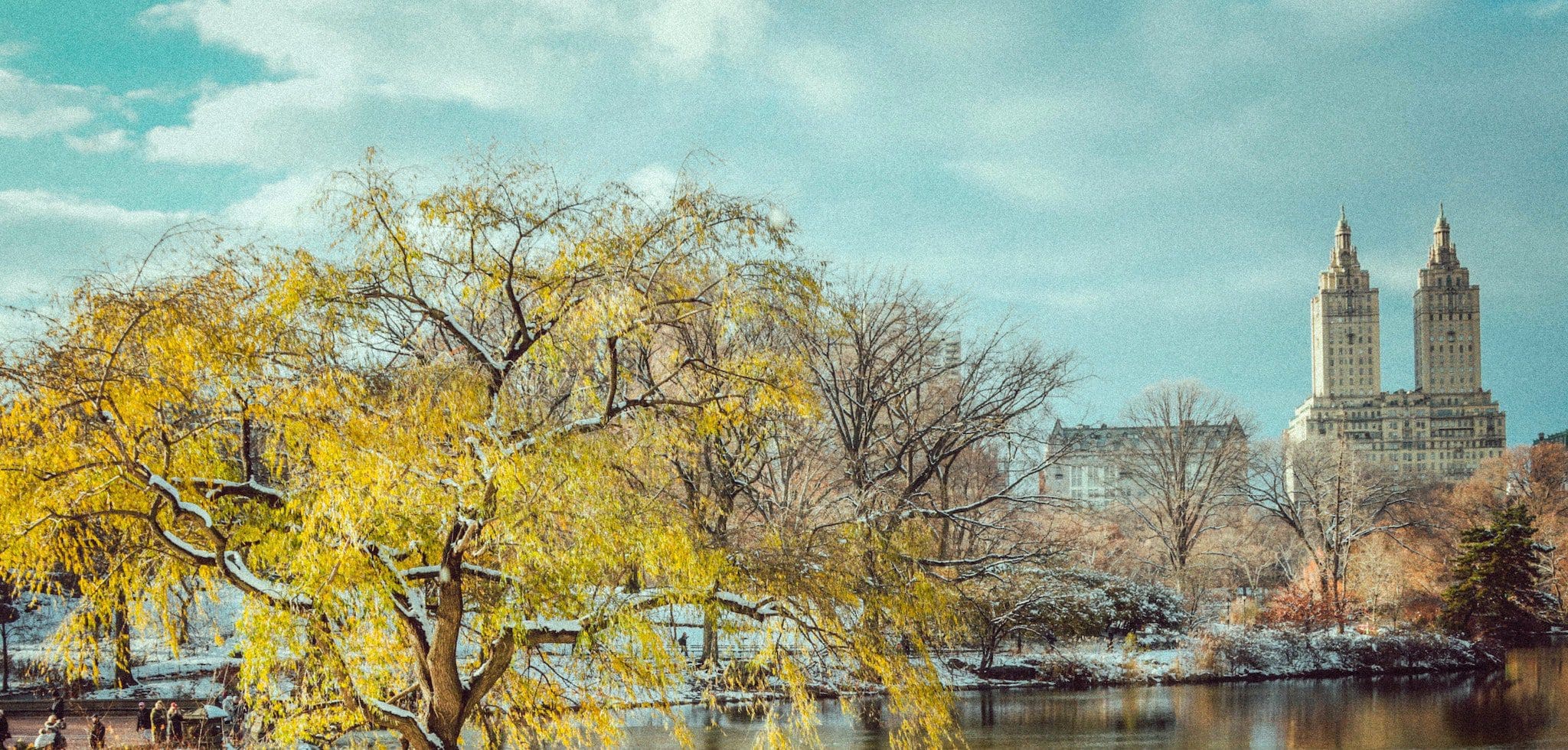 A serene landscape featuring a yellow-leaved tree by a pond, with light snow on the ground. In the background, two tall, twin-towered buildings rise against a partly cloudy sky. Sparse foliage and people walking are visible.