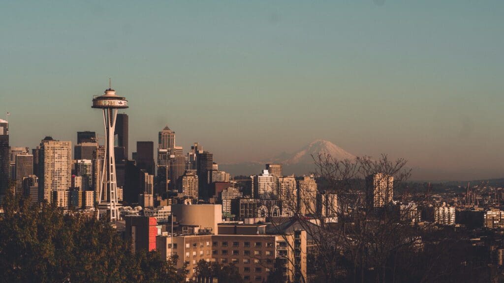 Seattle skyline at sunset, featuring the Space Needle and downtown buildings. Mount Rainier is visible in the background under a clear sky. Some trees are in the foreground, adding depth to the cityscape.