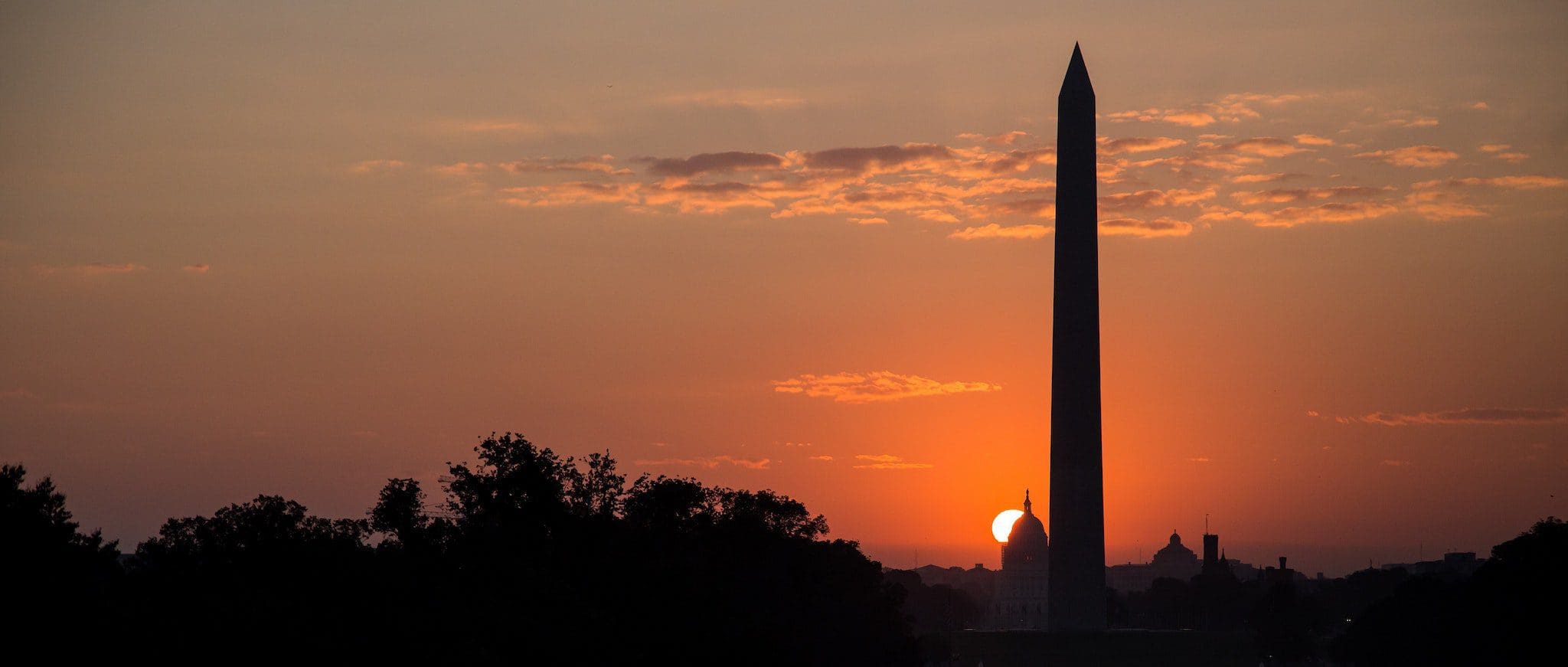 Silhouette of the Washington Monument against a vibrant sunset, with the sun partially hidden behind the monument. The sky is a mix of orange and purple hues, and trees are visible in the foreground.