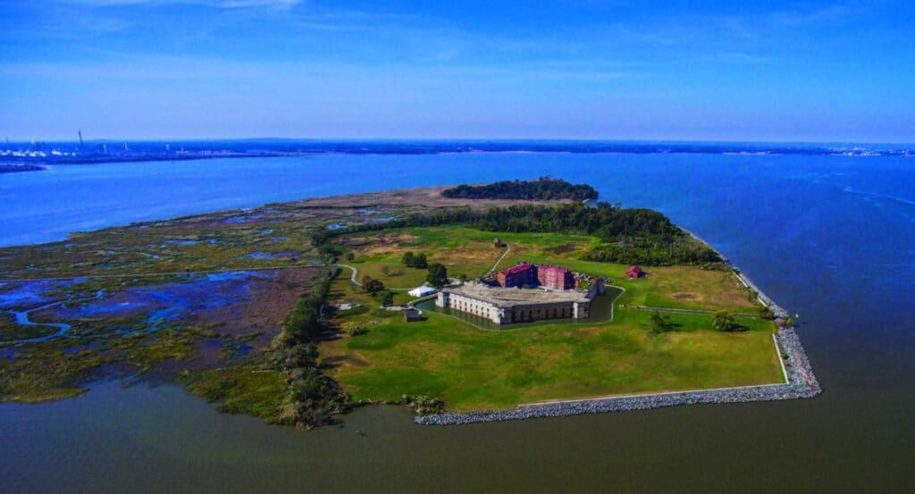 Aerial view of a small, grassy island surrounded by water, featuring a large historical fort with red and brown buildings. The island is bordered by rocky shores, and patches of greenery are visible. The horizon shows a blue sky with distant land.