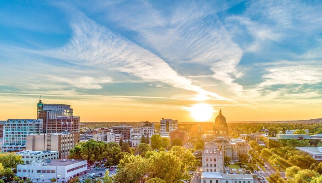 A cityscape at sunset featuring modern and historic buildings. The sun is setting on the horizon, casting a warm glow. A prominent dome-shaped building is silhouetted against a sky with streaked clouds. Greenery is visible in the foreground.