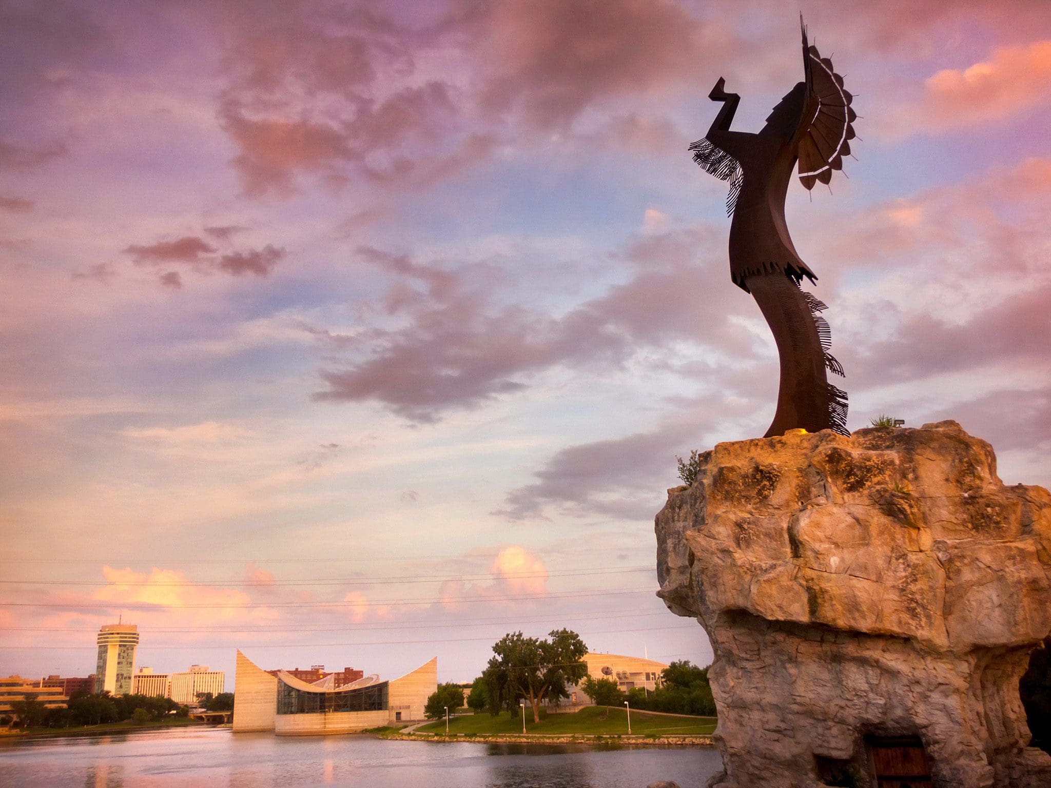 A towering metal sculpture of a Native American holding a feathered staff stands on a rocky pedestal by a river at sunset. The sky is painted in hues of pink and purple, and buildings are visible in the background.