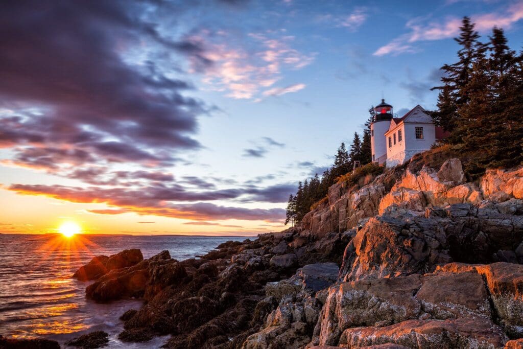 Sunset view of a lighthouse perched on a rocky coastline, surrounded by pine trees. The sky is a mix of pink, orange, and blue hues, with the sun setting over the ocean, casting a warm glow on the scene.