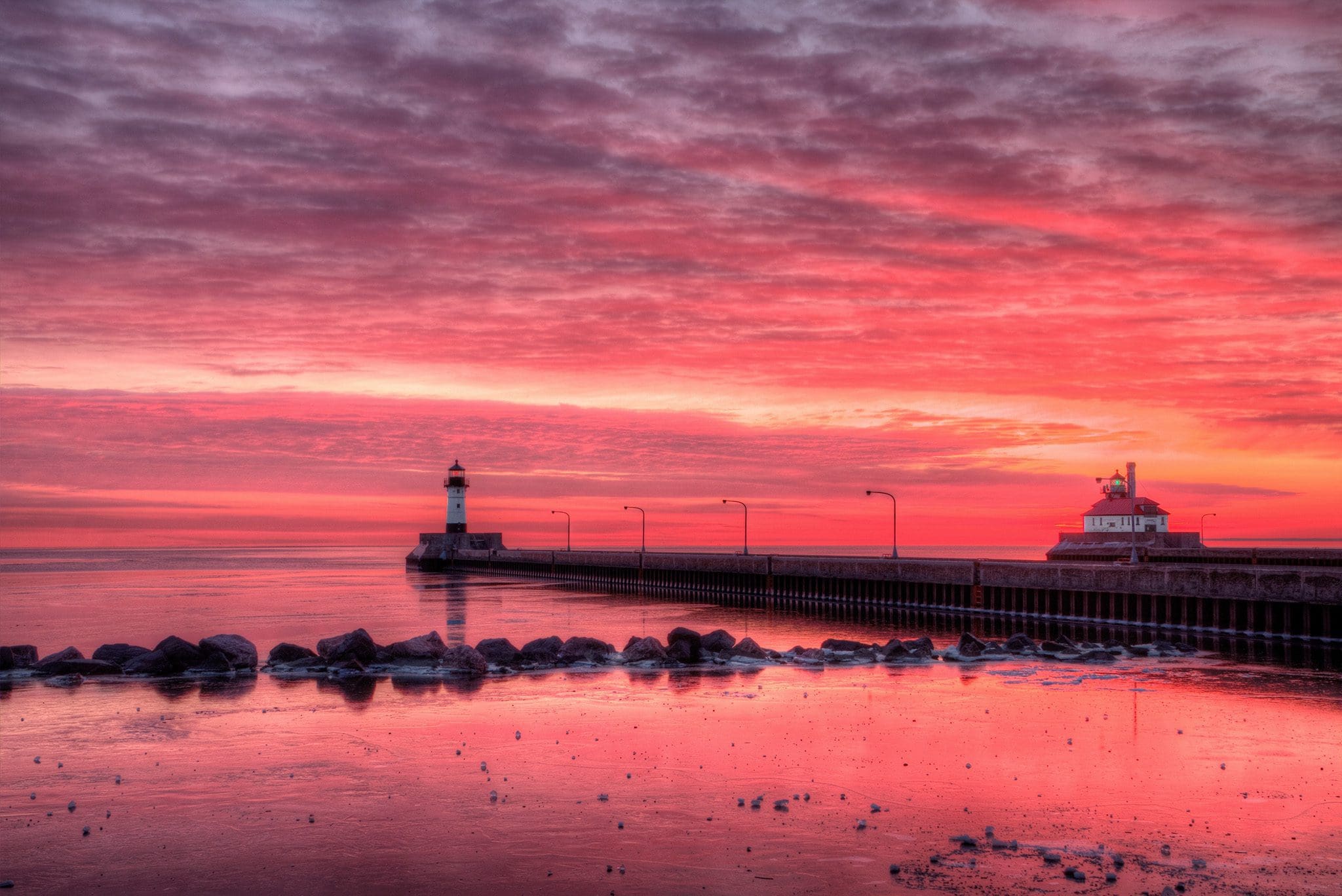 A vibrant sunset over a calm sea, with a sky filled with pink and purple hues. A lighthouse stands on the left pier, and a building is visible on the right. The water reflects the colorful sky, creating a serene and picturesque scene.