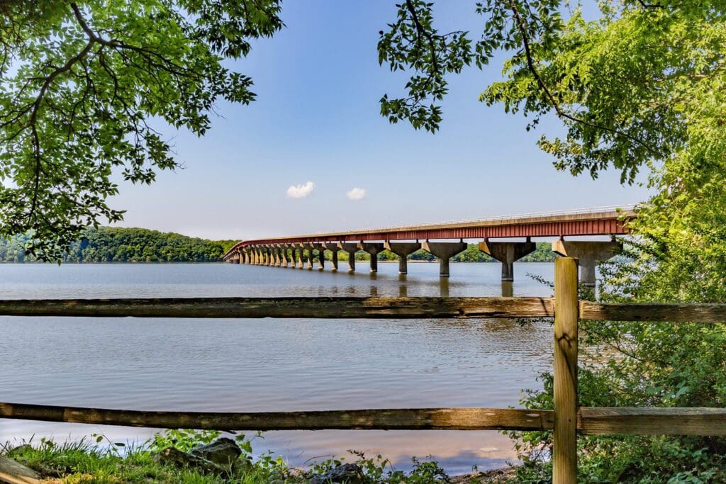 A long bridge with multiple supports extends across a calm body of water, framed by lush green trees and foliage. A wooden fence is visible in the foreground, adding depth to the scene under a clear blue sky with a few clouds.