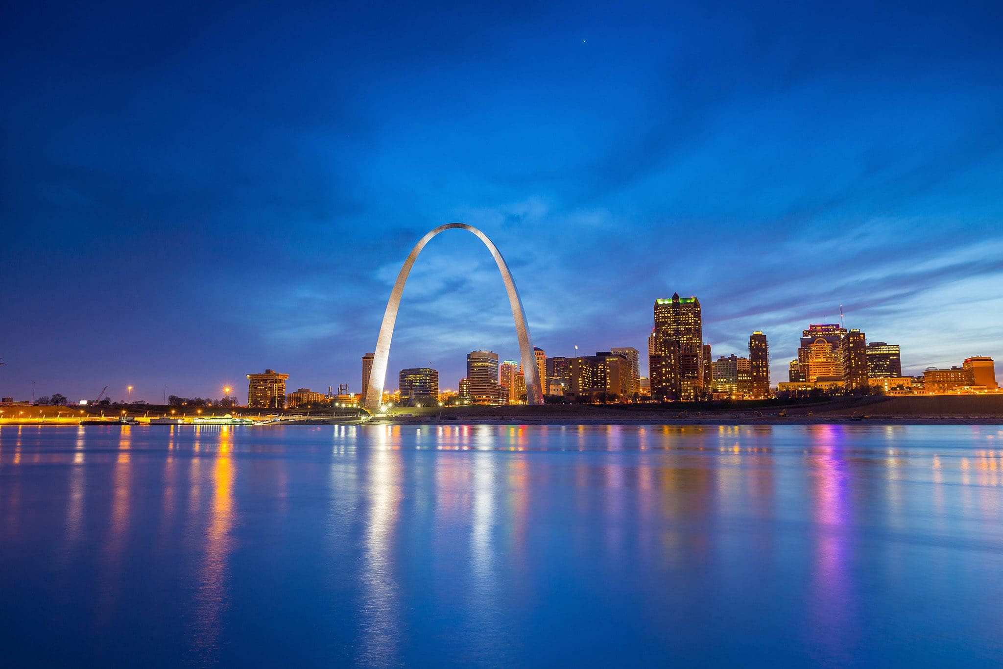 The image shows the St. Louis skyline at dusk, with the Gateway Arch prominently in the center. City lights illuminate the buildings, and their reflections shimmer in the calm waters of the Mississippi River.