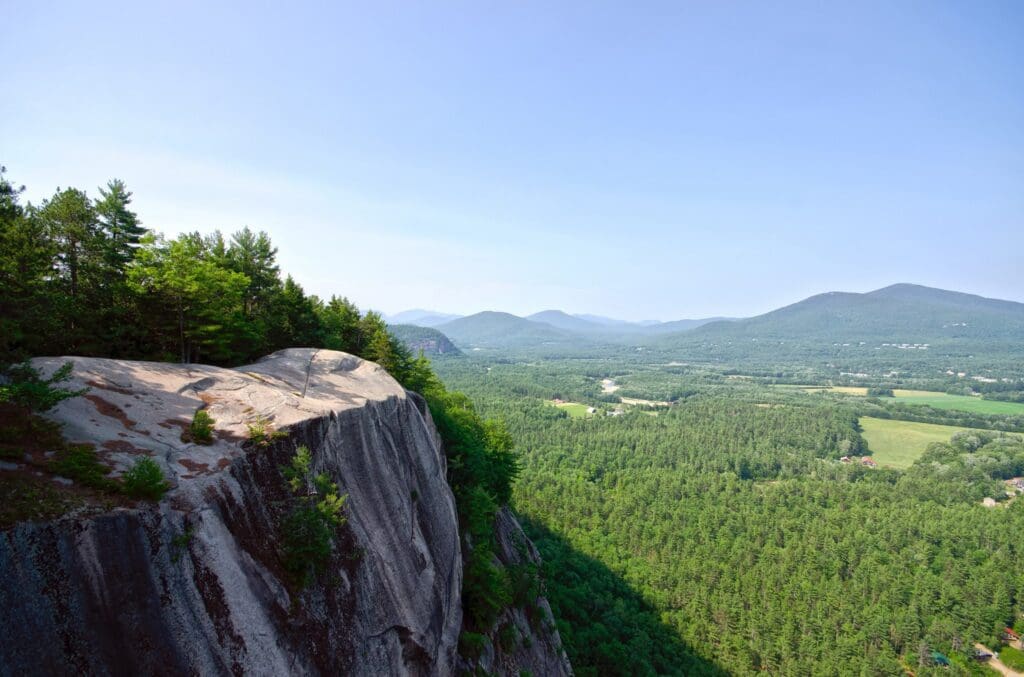 A scenic view from a rocky cliff edge surrounded by dense green forest. In the background, there are rolling hills and a clear blue sky, creating a serene, expansive landscape.